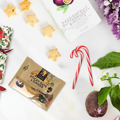 Packaged cookies with a focus on 'Sticky Date & Ginger' flavor, surrounded by decorative elements like candy canes and passion fruit.